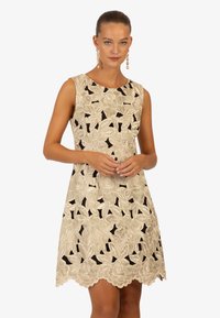 Mouwloze jurk van beige stof met bloemenborduursels en uitsparingen. Golfrand en een gladde textuur. Elegante uitstraling zonder extra accenten.