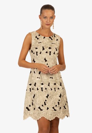 Mouwloze jurk van beige stof met bloemenborduursels en uitsparingen. Golfrand en een gladde textuur. Elegante uitstraling zonder extra accenten.