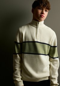 Pullover in pile color crema con texture a coste; presenta una striscia verde e blu attraversa il petto e un colletto alto con zip.