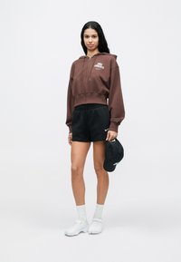 Νεαρή γυναίκα που φοράει καφέ φούτερ με φερμουάρ, μαύρο σορτς, κρατώντας μαύρο καπέλο, στέκεται με λευκές κάλτσες και αθλητικά παπούτσια σε λευκό φόντο.