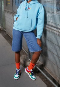 Sudadera azul claro con un bolsillo frontal y texto multicolor, combinada con pantalones cortos de mezclilla, calcetines naranjas y zapatillas multicolor. Fondo texturizado.