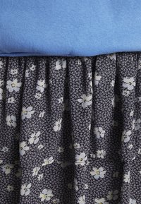 Top de algodão azul combinado com uma saia plissada de tecido escuro com um padrão floral branco sobre um fundo texturizado.