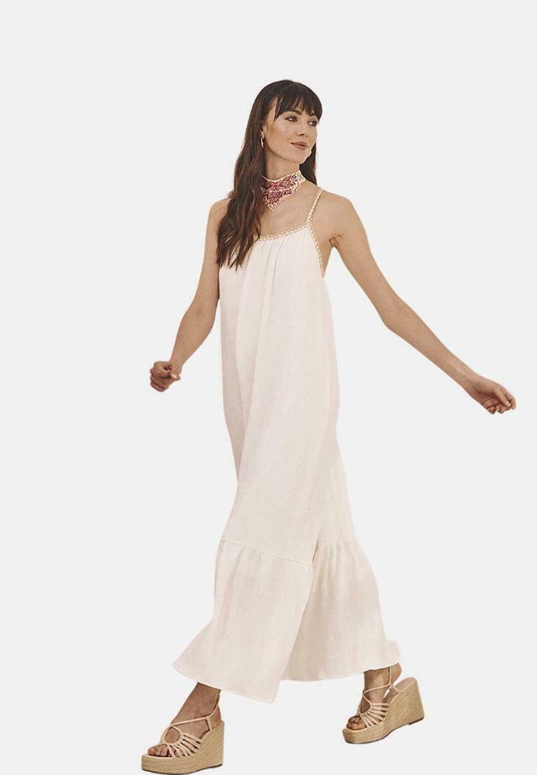Vestido maxi de tejido beige claro, con dobladillo acampanado, tirantes finos y una silueta holgada. La modelo lleva sandalias de cuña en color marrón.
