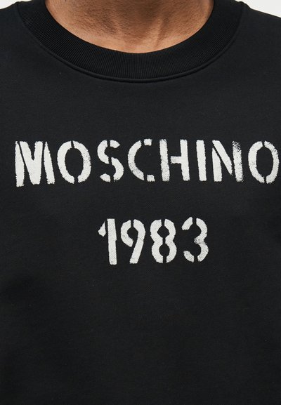 Μαύρο φούτερ με στρογγυλή λαιμόκοψη και λευκό φθαρμένο κείμενο "MOSCHINO 1983" στο στήθος, που φοριέται από ένα άτομο.