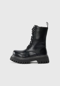 Juodos odinės kovinės boots su storu padu, priekyje su raišteliais, šone su užtrauktuku ir traukimo kilputė ant paprasto šviesaus fono.