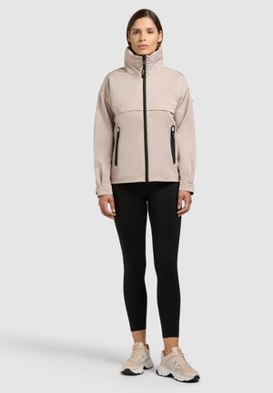 Frau steht und trägt eine beige Jacke mit hohem Kragen und schwarzen Reißverschlüssen, schwarze Leggings und beige Sneaker vor einem einfarbigen hellgrauen Hintergrund.