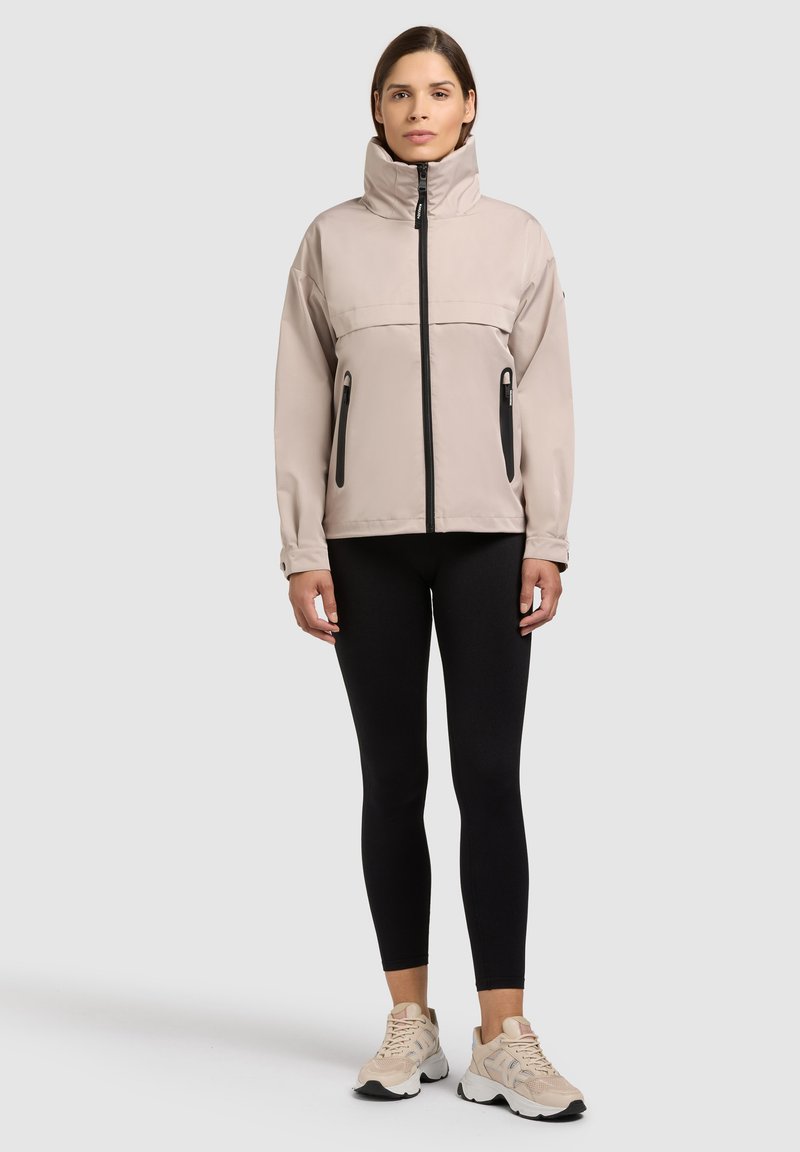 Femme debout portant une veste beige à col montant avec des fermetures éclair noires, des leggings noirs et des baskets beiges sur un fond gris clair uni.