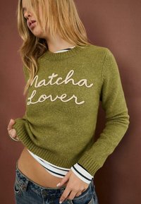 Oliven grøn sweater med "Matcha Lover" broderet i creme, med ribbede manchetter og kant, lagdelt over en stribet skjorte.