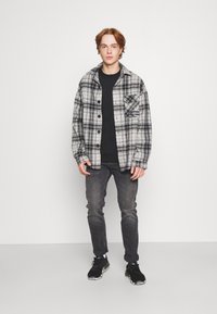 Chemise en flanelle à carreaux noir et blanc avec fermeture à boutons et poche avant, portée par-dessus un t-shirt noir ; associée à un jean foncé et des baskets noires.