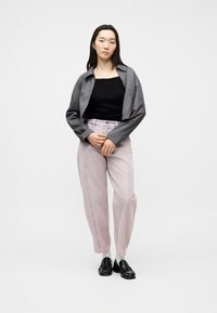 Νεαρή γυναίκα που στέκεται με τα χέρια δεμένα, φορώντας γκρι σακάκι, μαύρο τοπ, ανοιχτό ροζ παντελόνι, άσπρες κάλτσες και μαύρα μοκασίνια, σε λευκό φόντο.