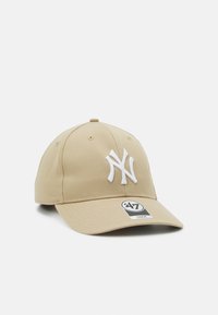 Tan baseball cap with a curved brim, featuring a white embroidered New York emblem on the front and a logo tag on the side. Cotton material.