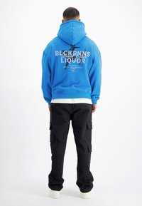 Blauwe hooded sweatshirt met een grafische print op de achterkant met de woorden "BLCHBNNS Limited Edition LIQUOR." Gecombineerd met zwarte cargobroeken.