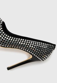 Black high-heeled shoe with a pointed toe, covered in silver studs, featuring a sleek silhouette and a smooth leather finish.
