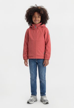 Dieťa s kučeravými vlasmi, ktoré má na sebe koralovú bundu s kapucňou, modré džínsy a sivé tenisky, stojí a usmieva sa na jednoduchom pozadí.