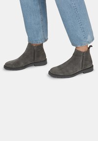 Stivaletti alla caviglia in suede grigio con cerniera laterale, suola in gomma nera e una texture liscia, abbinati a jeans azzurri arrotolati.