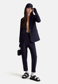 Ciemnoniebieski garnitur w paski z dopasowaną marynarką i zwężanymi spodniami, w połączeniu z czapką w kolorze czarnym, usztywnioną torebką oraz masywnymi sandałami.