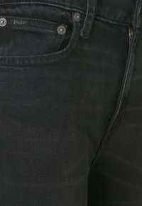 Svarta denim jeans med synlig sömnad, en framficka med en etikett märkt "Polo" och silverfärgade knappar. Tyget har en svag lyster.