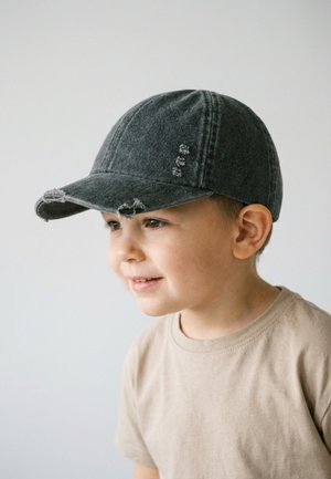 Junger Junge mit hellem Teint, trägt eine abgenutzte schwarze Baseballkappe und ein beiges T-Shirt, lächelt und schaut leicht nach rechts vor einem schlichten Hintergrund.