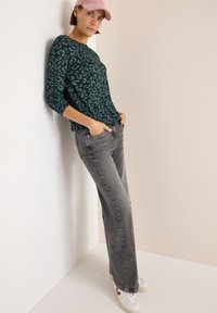 Langärmliges, teal-farbenes Oberteil mit einem schwarzen Leopardenmuster, kombiniert mit grauen, geraden Jeans und weißen Turnschuhen, vor einem neutralen Hintergrund.