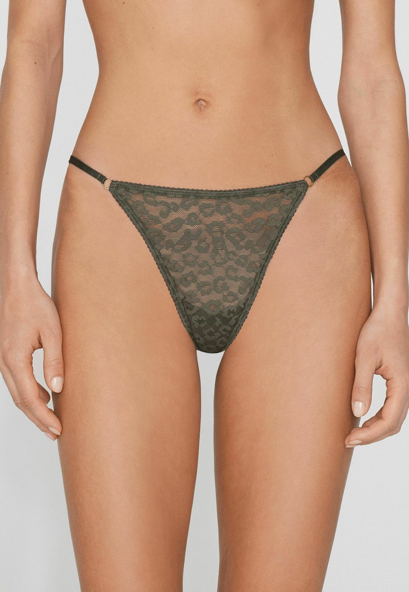 Femme portant un string léopard transparent vert avec des fines lanières sur les côtés, sur un fond uni.