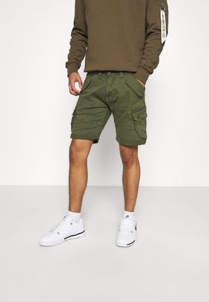 Mężczyzna ubrany w oliwkowo-zielone szorty cargo, brązową bluzę z długim rękawem z naszywką na rękawie, białe skarpetki i białe sneakersy Nike stoi na tle jednolitego tła.