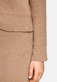 Nahaufnahme einer Hand, die neben einem beigen, strukturierten Strickoutfit mit goldenen Fadenakzenten und einem kleinen runden Metallknopf-Detail ruht.