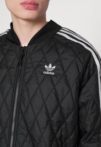 Černá prošívaná bomber bunda se zipem, žebrovaným límcem a bílými pruhy na rukávech. Logo Adidas zobrazené na hrudi.