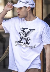 Camiseta blanca de algodón con un gráfico negro de un hombre a caballo alado y la letra "K". Combinada con una gorra blanca que lleva "Gianni Kavanagh".