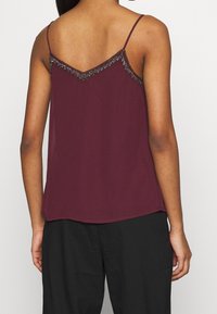 Femme portant un haut sans manches bordeaux avec des détails en perles et des fines bretelles, associé à un pantalon noir ample, vu de dos.