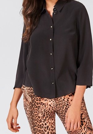 Vrouw die een zwart blouse met knopen en driekwart mouwen draagt, gecombineerd met een broek met luipaardprint, tegen een effen achtergrond.