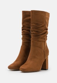 Stivali in suede marrone a caduta con tacco spesso, caratterizzati da una texture liscia e un design arricciato lungo il gambale per un tocco di dettaglio in più.