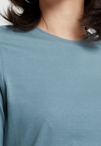 Camiseta de rayas azul claro con cuello redondo. Detalles de costura discretos y textura suave, parcialmente visible bajo el cabello oscuro.