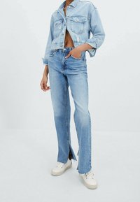 Chaqueta de mezclilla de color azul claro con bolsillos frontales, de diseño corto; combinada con jeans de color azul claro que presentan aberturas laterales. Zapatillas blancas completan el atuendo.