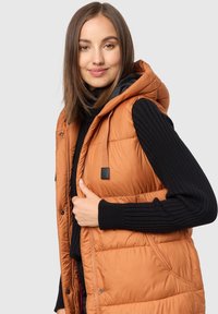 Vadderad ärmlös puffervest i varm orange med huva, med dragkedja och sidofickor. Texturerad svart stickad långärmad tröja under.