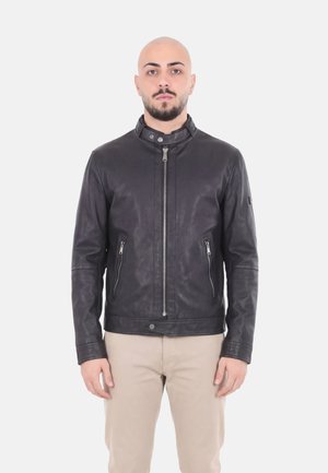 Uomo con testa rasata e barba che indossa una giacca di pelle nera e pantaloni beige, in piedi su sfondo bianco.