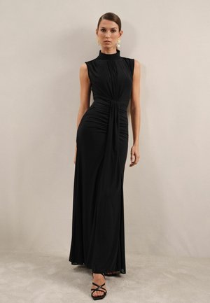 Schwarzes ärmelloses Maxikleid mit hohem Kragen, Raffung, fließendem Stoff und schmalem Schnitt, kombiniert mit schwarzen Sandalen mit Riemchen.