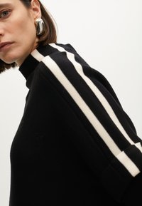 Černý svetr s vysokým límcem, má tři bílé pruhy podél rukávů. Hladká textura, přiléhavý střih a ohrnuté rukávy.