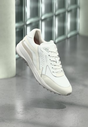 Pantof sportiv alb și bej cu șireturi, guler căptușit și talpă texturată, poziționat în aer deasupra unui podea din beton lustruit.