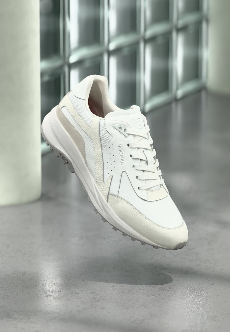 Baskets athlétiques blanches et beiges avec lacets, col rembourré et semelle texturée, positionnées en suspension au-dessus d'un sol en béton poli.