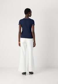 Chemise bleu marine à manches courtes avec des bordures blanches et rouges, assortie à un pantalon large blanc et des talons noirs, vue de dos.