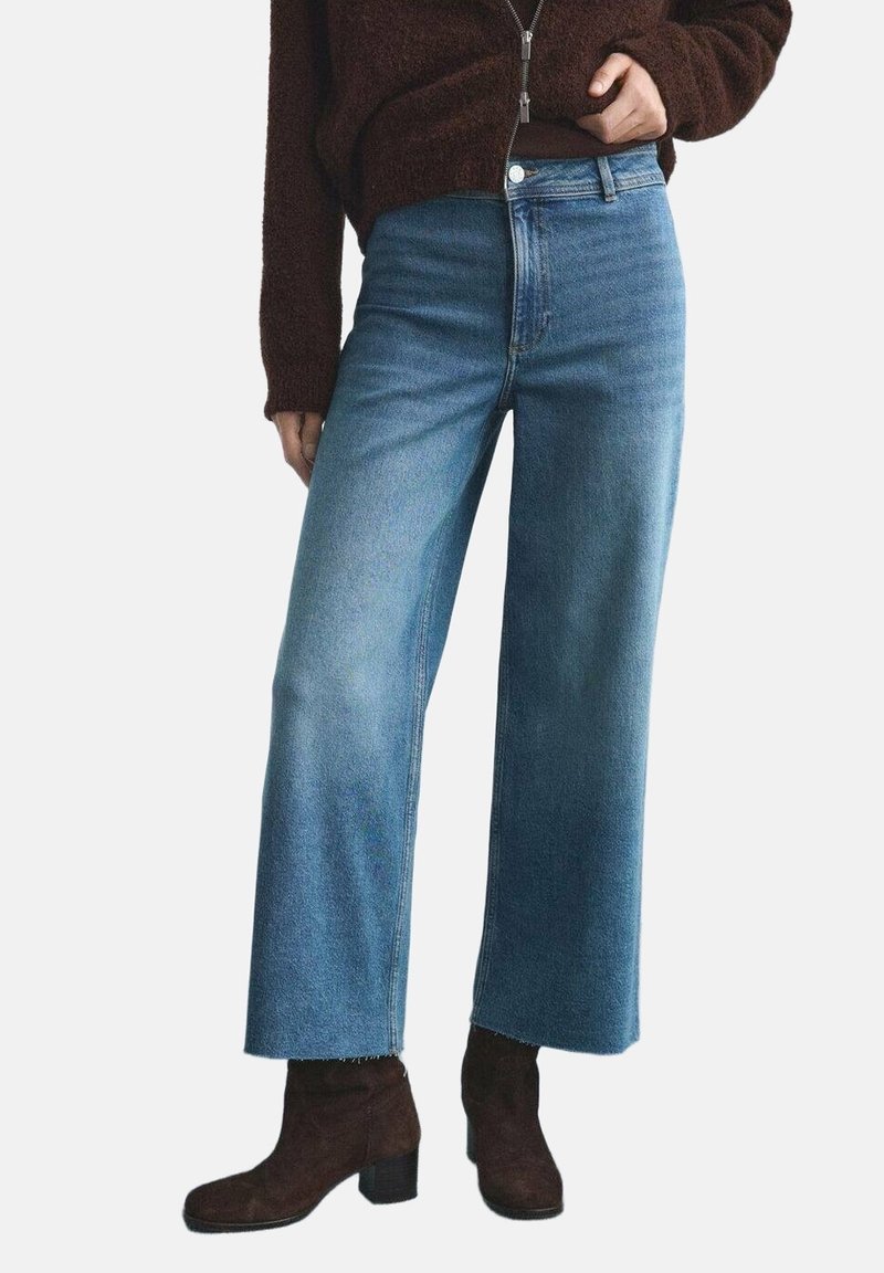 Une personne porte un jean large bleu clair associé à des bottines marron foncé et un pull zippé marron foncé, debout devant un fond uni.