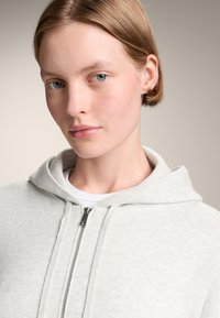 Svijetlo siva zip-up dukserica s mekanom teksturom, koja ima kapuljaču i srebrni patentni zatvarač. Nošena preko bijele majice, bez istaknutih uzoraka ili akcenta.