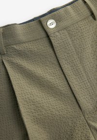 Olive green trousers with a textured surface, pleated front, and a single button closure. Features a contrasting dark interior waistband.