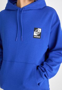 Blå hoodie gjord av mjukt tyg, med en framficka, dragsko-huva och en logotyp-patch på vänster sida.