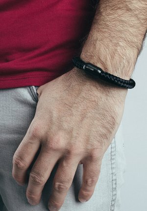 Black braided bracelet with a sleek black clasp, worn on a male wrist, featuring a textured surface and visible stitching details.