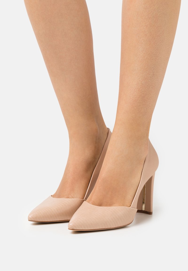 Jambes féminines portant des escarpins couleur nude à bout pointu avec talons blocs sur un fond uni.