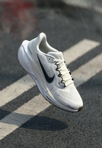 Bijela atletska cipela s mrežastim gornjištem, crnim swoosh logotipom i teksturiranim potplatom. Ima vezanje na vezice i vidljive akcentne udobnosti.