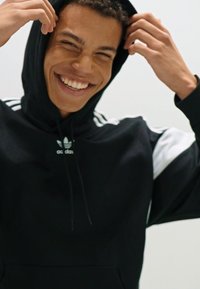 Felpa nera con accenti e strisce bianche. Presenta un cappuccio con cordino e tasca frontale. Il materiale sembra morbido con una texture liscia.