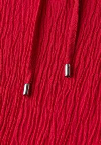 Tessuto rosso con texture, presenta pieghe verticali e due cordini che terminano con punte metalliche argentate. Il materiale appare leggero e flessibile.