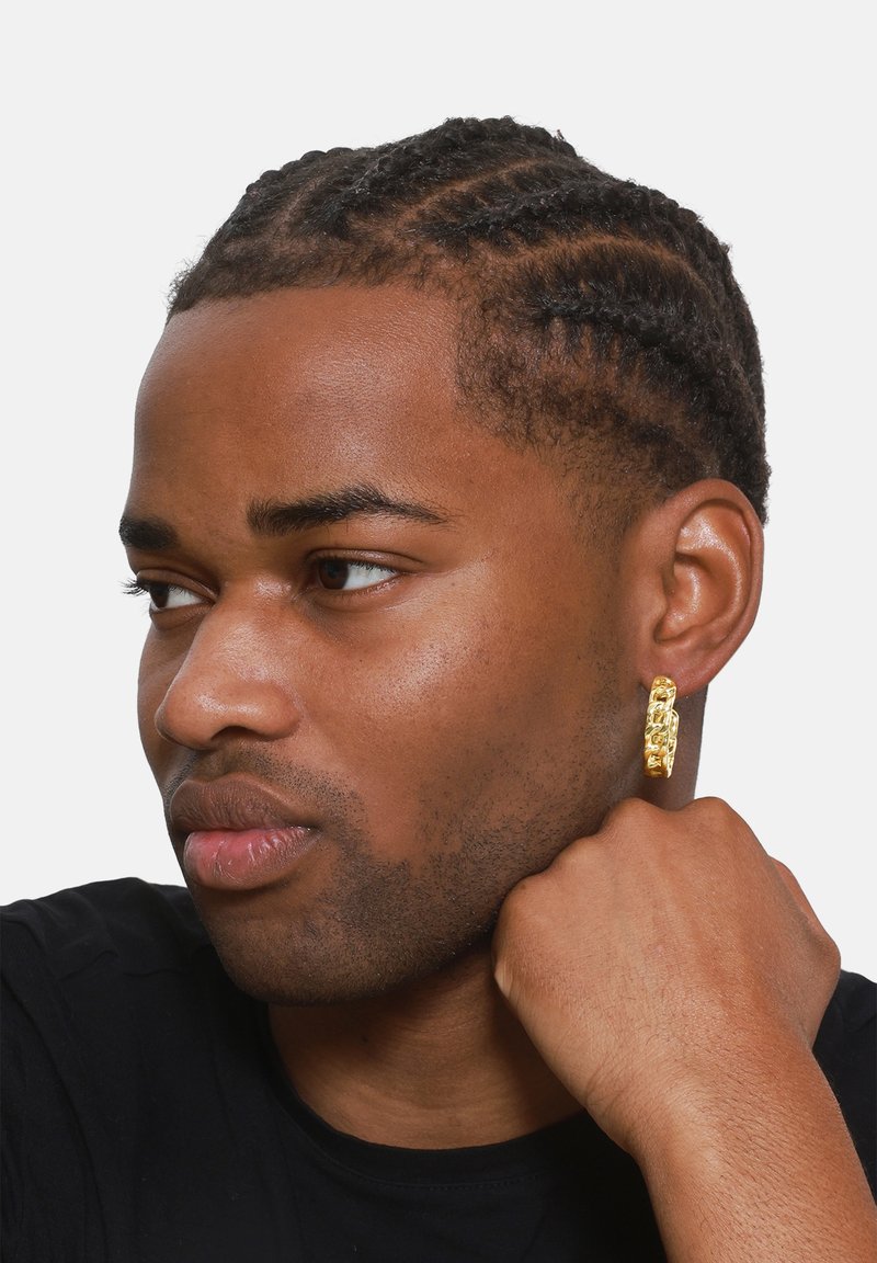 Boucle d'oreille en or avec un design en chaînes texturées, portée par une personne aux cheveux tressés, sur fond blanc uni.